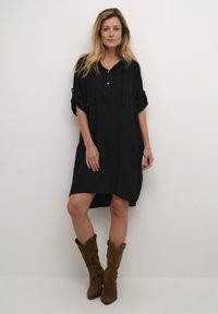 Rochie din in negru cu glugă, închidere cu nasturi, mâneci răsucite și slituri laterale. Asortată cu cizme din suede maro până la genunchi.