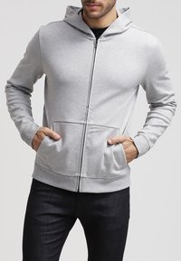 Sudadera gris con cremallera, confeccionada en una suave mezcla de algodón, con un bolsillo tipo canguro y puños acanalados. Presenta una capucha con cordón y una textura de tejido suave.