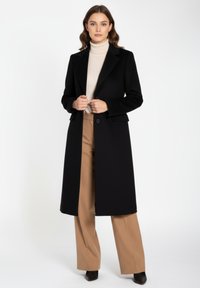Black, long coat with a structured design, notched lapels, two front pockets, worn over a beige turtleneck and tan wide-leg trousers.