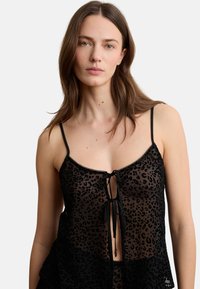 Frau mit langen braunen Haaren, die ein schwarzes, transparentes Oberteil mit Leopardenmuster, dünnen Trägern und vorderer Schnürung vor einem schlichten Hintergrund trägt.