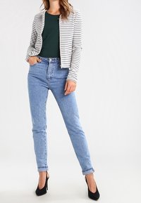 Randig vit och grå cardigan över en grön topp, kombinerad med ljusblå högmidjade jeans och svarta klackar. Släta material och figursydd design.