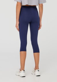Leggings capri blu navy con una texture liscia, vita alta e design aderente. Indossati con un top corto grigio chiaro e scarpe da ginnastica bianche.
