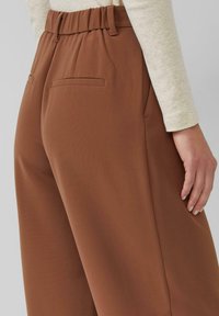 Brązowe spodnie z elastycznym paskiem w talii, bocznymi kieszeniami i gładką fakturą. Tkanina lejąca się luźno, o czystym, prostym kroju.