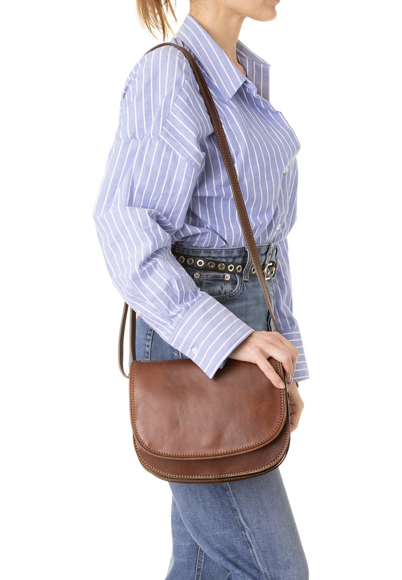 Borsa a tracolla in pelle marrone con patta arrotondata, cuciture decorative e una singola tracolla regolabile, indossata sopra una camicia a righe blu e bianche.