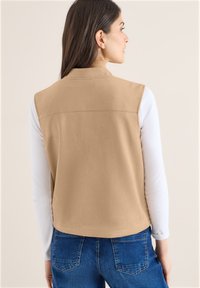 Femme aux cheveux noirs et lisses portant un gilet beige sans manches par-dessus une chemise blanche à manches longues et un jean bleu, dos tourné à la caméra.