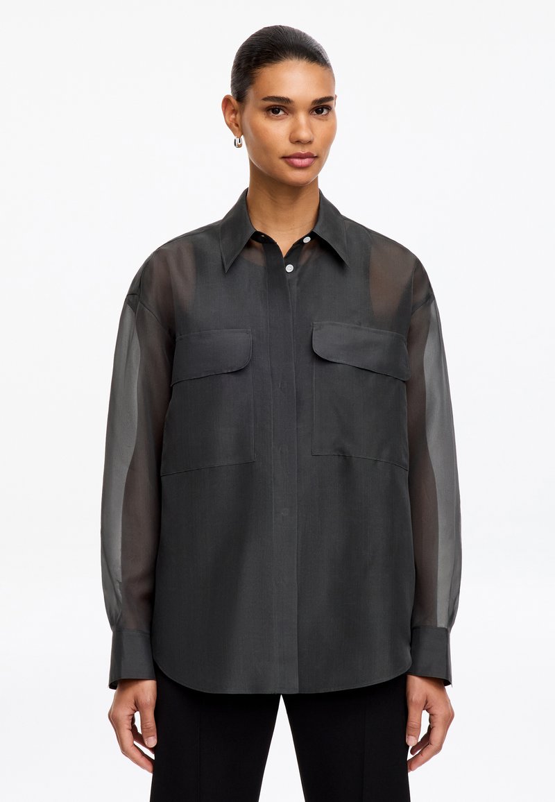 Femme portant une chemise longue noire transparente avec poches poitrine et col, debout devant un fond blanc uni.