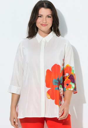 Frau mit dunklem Haar trägt eine weiße Bluse mit Knöpfen, die auf einer Seite ein großes rotes und mehrfarbiges Blumenmuster aufweist, kombiniert mit roten Hosen.