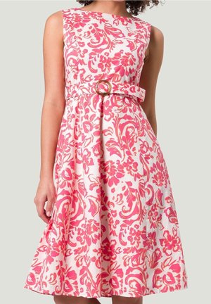 Robe blanche sans manches avec motif floral rose et ceinture assortie avec boucle ronde, portée par une personne sur fond clair uni.