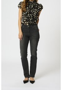 Svart blus med volanger och beige blommönster, i kombination med åtsittande mörka jeans och svarta spetsiga klackar, vilket visar en avslappnad outfit.