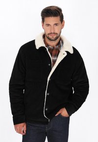 Sort kordfløjljakke med krave i hvid faux shearling, to forlommer og metalknapper, båret over enternet skjorte.