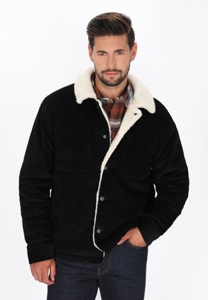 Schwarze Cordjacke mit einem weißen Kunstpelzkragen, zwei Vordertaschen und Metallknöpfen, getragen über einem karierten Hemd.