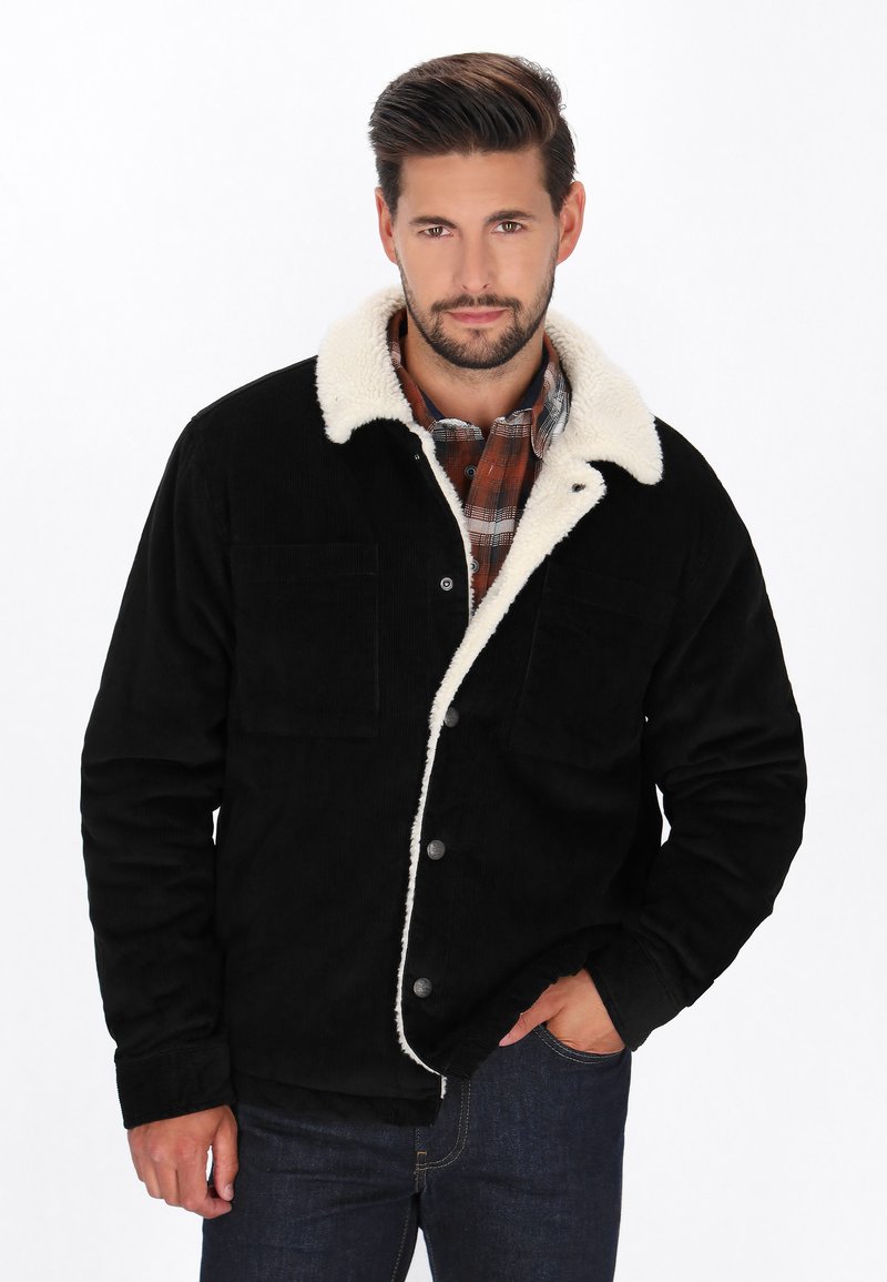 Sort kordfløjljakke med krave i hvid faux shearling, to forlommer og metalknapper, båret over enternet skjorte.