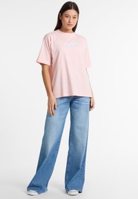 Lys rosa oversized t-shirt med korte ærmer og blå logo-tekst på brystet, parret med falmede blå vide jeans og hvide sneakers.