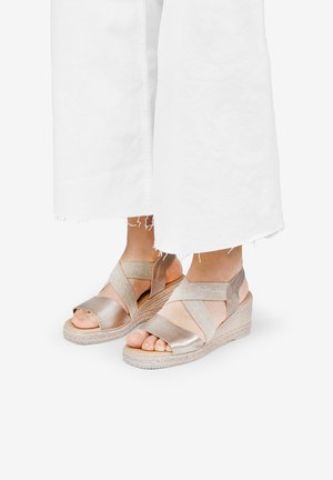Sandalias de cuña metálicas doradas con tiras de tela entrecruzadas y dedos al aire, que cuentan con una suela de yute trenzado y detalles accentuados texturizados.