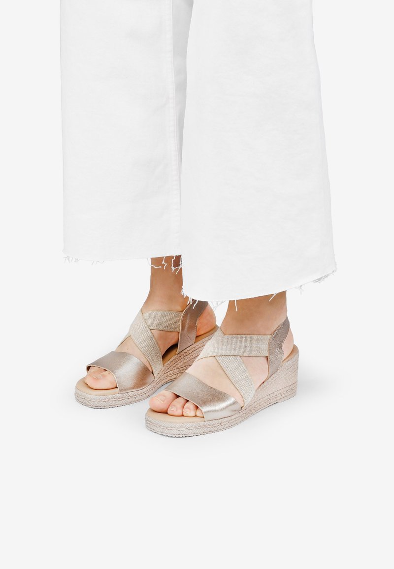 Sandalias de cuña metálicas doradas con tiras de tela entrecruzadas y dedos al aire, que cuentan con una suela de yute trenzado y detalles accentuados texturizados.