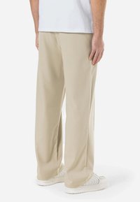 Pantalons larges beiges en tissu doux, avec une coupe décontractée et des fentes sur les côtés. Portés avec des baskets blanches, mettant en avant un design simple.