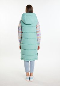 Gilet lungo imbottito di un verde menta, dotato di cappuccio, cuciture orizzontali e spacchi laterali. Indossato sopra un maglione colorato con motivo e jeans chiari.