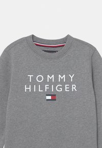Sudadera gris con cuello redondo, que presenta el texto "TOMMY HILFIGER" en blanco. Incluye un pequeño parche de logo en rojo, blanco y azul. Textura suave.