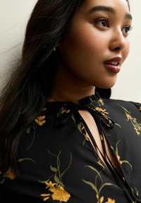 Schwarze Bluse mit gelben Blumen, mit einer Krawatten-Halslinie und einem Schlüsselloch-Ausschnitt, glattem Stoff und langen Ärmeln mit gerafftem Detail.