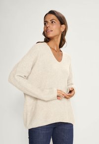 Crème-kleurige gebreide trui met een V-hals, verlaagde schouders en lange mouwen. Heeft een losse pasvorm en ribbelstructuur.