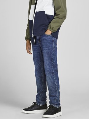 Jean en denim bleu avec une coupe droite, associé à une veste en block coloré comportant des sections vertes, blanches et marines. Baskets noires.