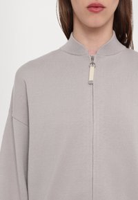 Ljusgrå zip-up tröja med hög krage, ribbad textur och en kontrasterande dragkedja, med en avslappnad passform och sänkt axel.