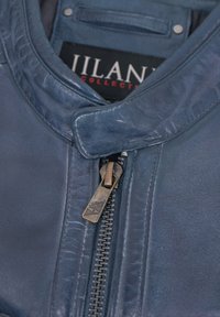 Blaue Lederjacke mit Stehkragen, ausgestattet mit einem Metall-Reißverschluss und einem sichtbaren Etikett "Jilani Collection" im Inneren. Glatte Textur.