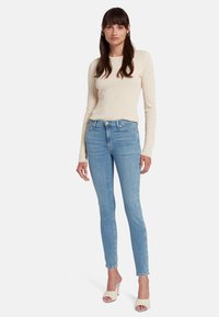 Maglia a maniche lunghe di colore crema con texture, abbinata a jeans skinny azzurri e mules open-toe, evidenziando uno stile casual e aderente.