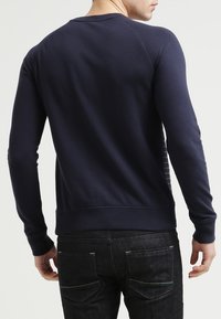 Hombre con una camisa de manga larga azul marino ajustada y jeans negros, mostrado de espaldas contra un fondo claro y liso.