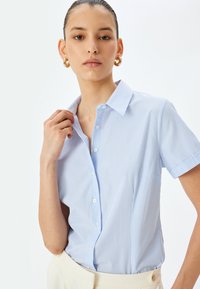Lichtblauwe, kortaarmige shirt met verticale witte strepen, knoopsluiting aan de voorkant en een enkele borstzak. Gladde stof met een kraag.
