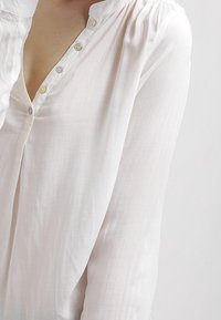 Vit, lättviktig skjorta i tyg med långa ärmar, en placket-halsringning med små knappar och en mjuk, slät textur.