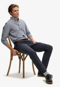 Overhemd met knopen in een marineblauw en wit geruite patroon, gecombineerd met donkere chino's, grijze sokken en donkere bootsschoenen. Een houten stoel is zichtbaar.