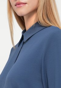 Gros plan d'une personne aux longs cheveux blonds portant une chemise bleue à col, montrant les lèvres, le menton et le cou sur un fond blanc.