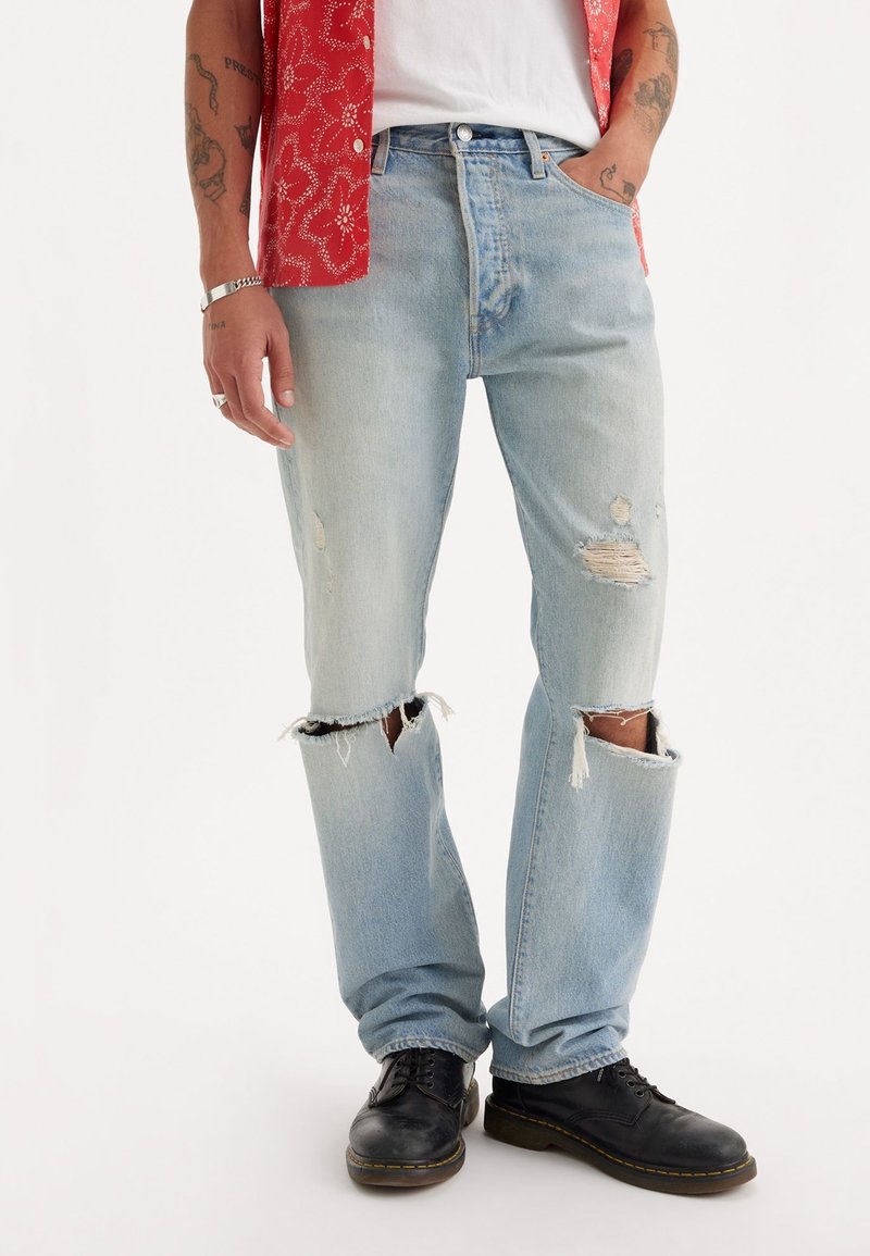 Jeans de mezclilla azul claro desgastados con corte recto, dobladillo deshilachado y detalles rasgados. Combinados con zapatos negros y una camisa roja con estampado floral.