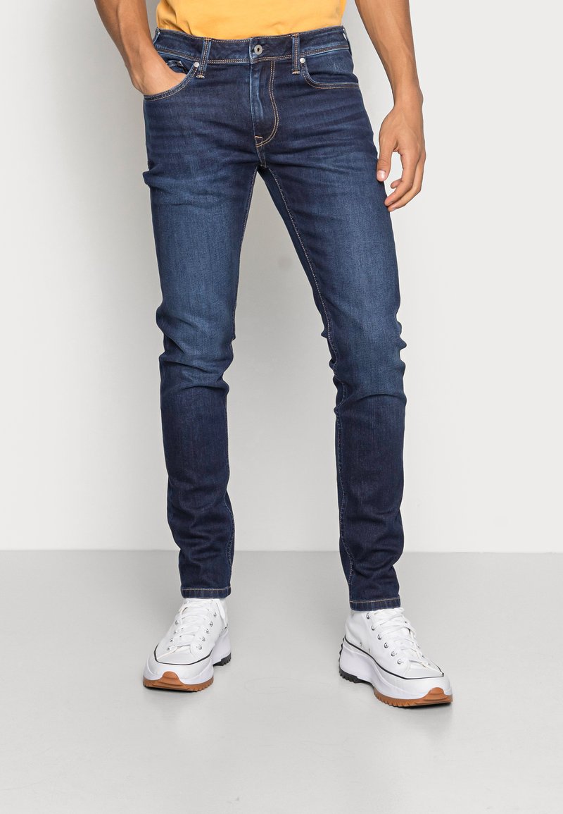 Jeans de ganga azul escura com um corte slim, design de cinco bolsos, costura visível e cintura média. Combinados com ténis brancos.