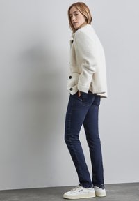 Texturerad vit ullblandad jacka med svarta knappar, i kombination med mörkblå slim-fit jeans och vita sneakers. En enkel, modern design.