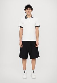 Camisa polo branca com colarinho preto e padrão de zebra, detalhes nas mangas em preto, calções pretos e ténis brancos. Modelo está contra um fundo simples.