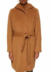 Vrouw in een camel-kleurige wollen jas met een brede kraag en een riem die om de taille is gebonden, weergegeven van schouders tot midden boven de dij.