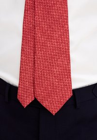Röd mönstrad slips av texturerad tyg med en geometrisk design. Slipsen har en klassisk spetsig ände och bärs över en vit skjorta och mörka byxor.