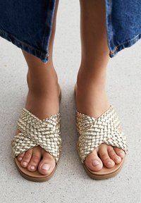 Kultaiset kudotut sandaalit, joissa on ristikkäinen muotoilu, tasapohja ja avoin varvas. Likainen tekstuuri ja neutraalin sävyinen pohja täydentävät kudontaa.