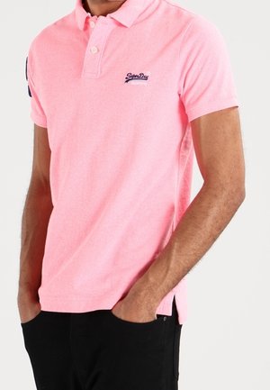 Homme portant un polo à manches courtes rose vif avec un logo brodé sur la poitrine, les mains dans les poches, devant un fond clair uni.