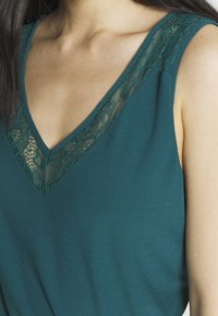 Top verde acqua con scollatura a V caratterizzato da dettagli in pizzo lungo i bordi. Realizzato in un tessuto liscio, ha una vestibilità morbida e design senza maniche.