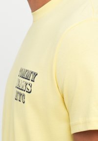 Geltona medvilninė marškinė su ryškiu atspaudo logotipu "TOMMY JEANS NYC" tamsiomis spalvomis. Standartinis apvalus kaklas ir trumpi rankovės.