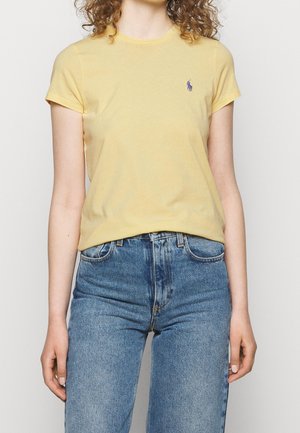 Kvinde med krøllet hår iført en gul t-shirt med korte ærmer og blå højtaljede jeans, der står foran en ensfarvet baggrund.