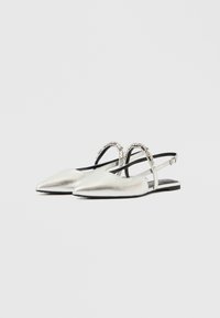 Μεταλλικά ασημί slingback μπαλαρίνες με μυτερή μύτη και διακοσμητικό λουράκι με τρουκς στο τακούνι. Λείο υλικό και χαμηλό μαύρο τακούνι.
