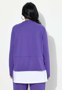 Maglia viola a maniche lunghe con un design a strati, caratterizzata da una texture liscia e un sottostrato bianco visibile all'orlo. Vestibilità semplice e rilassata.