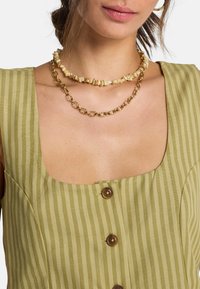 Πράσινο φόρεμα με ρίγες, τετράγωνο ντεκολτέ και λεπτομέρεια με καφέ κουμπιά, συνδυασμένο με δύο κολιέ: μια χρυσή αλυσίδα και ένα κολιέ με φυσικές χάντρες.