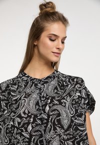 Sort bluse med paisley mønster i hvid, rund halsudskæring og korte ærmer, lavet af et let materiale.
