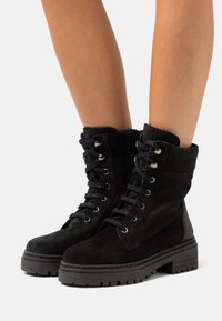 Botas de tornozelo em camurça preta com atacadores na frente, detalhes em metal prateado e sola de borracha grossa. As botas apresentam um fecho com zipper e detalhes texturizados.