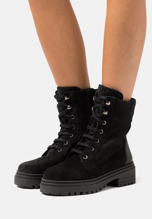 Botas de tornozelo em camurça preta com atacadores na frente, detalhes em metal prateado e sola de borracha grossa. As botas apresentam um fecho com zipper e detalhes texturizados.